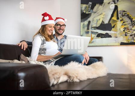Femme enceinte et mari portant un chapeau de père Noël et faisant du shopping sur un ordinateur portable, Munich, Allemagne Banque D'Images