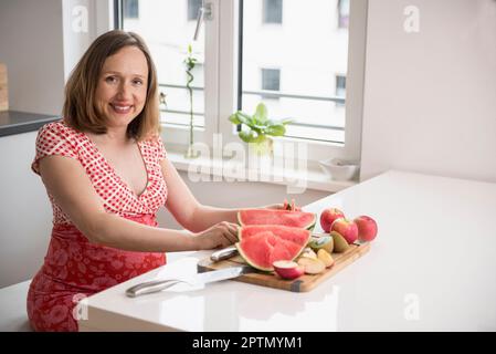 Femme enceinte coupant des tranches de pastèque dans la cuisine, Munich, Bavière, Allemagne Banque D'Images