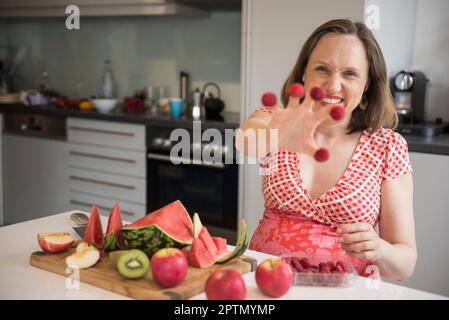 Femme enceinte montrant des framboises sur ses doigts dans la cuisine et souriante, Munich, Bavière, Allemagne Banque D'Images