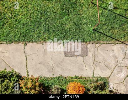 Ancienne chaussée fissurée avec pelouse verte et de petits arbustes autour. Vue de dessus, avec balcon en étage élevé à plat. Banque D'Images