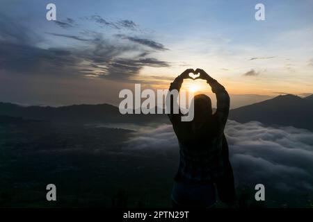 Silhouette d'une jeune femme doigts formant un coeur à l'aube, Uluwatu, Bali, Indonésie Banque D'Images