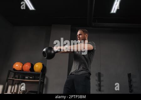 Athlète musclé qui fait des balançoires de kettlebell au gymnase crossfit, tenue sur fond gris de gym avec équipement. Haltérophilie faire de l'exercice à l'intérieur, faire de l'exercice. Échauffement de l'instructeur CrossFit Banque D'Images