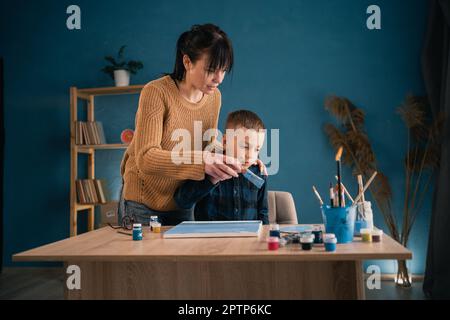 une jeune mère heureuse aide son fils à peindre de la peinture acrylique sur toile à la maison. concept de passe-temps commun. Banque D'Images