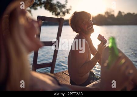 Jeune homme heureux mangeant de la pastèque sur un quai au bord de l'eau pendant le coucher du soleil d'été Banque D'Images