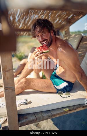 Une vue rapprochée d'un jeune homme pose pour la photo tout en étant assis à l'ombre sur le lit de soleil à la plage et en appréciant la pastèque pendant un bel été Banque D'Images