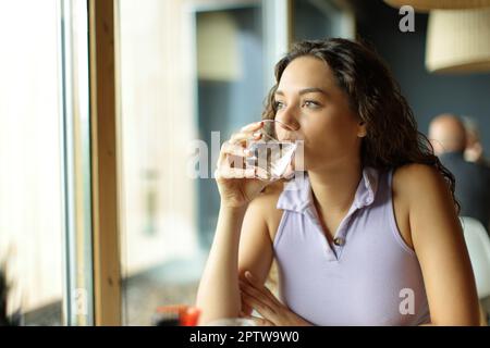 Une femme distrait de l'eau potable dans un restaurant en regardant loin par une fenêtre Banque D'Images