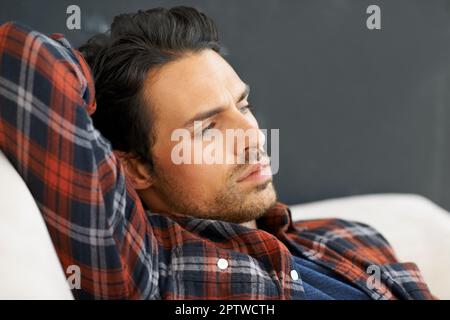 Sensation de copain et de mauvaise humeur. Un magnifique jeune homme allongé sur un canapé avec le bras derrière sa tête et regardant dans la distance avec une expression de contemplati Banque D'Images