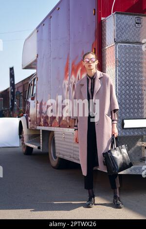 Jeune fille dans un manteau non attaché se tient près d'un camion rouge, profitant du soleil. Miroir de lunettes de soleil avec la rue réfléchissant dedans. Banque D'Images