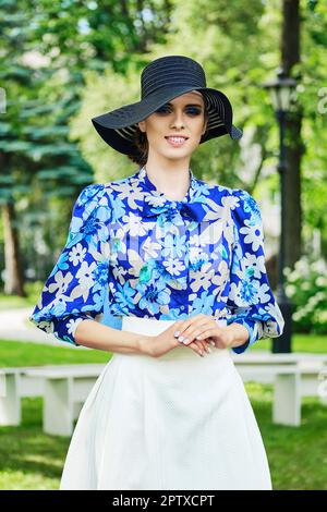 Portrait extérieur de fille souriante en chapeau noir à large bord, chemisier bleu et jupe blanche. Banque D'Images