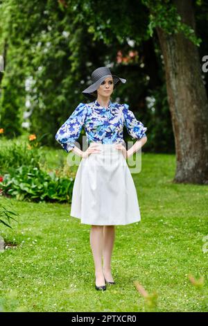 Portrait d'une fille souriante en chapeau noir à large bord, blouse bleue et jupe blanche debout sur l'herbe dans le parc. Banque D'Images