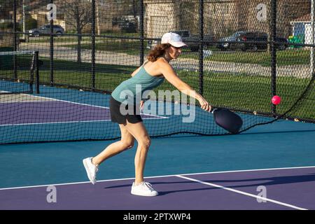 Un joueur de pickleball féminin retourne une balle rose vif à la ligne de base sur un terrain dédié dans un parc public. Banque D'Images