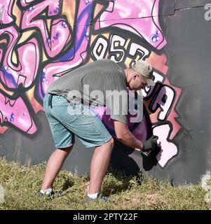 Jeune artiste de graffiti caucasien de sexe masculin dessinant de grandes peintures d'art de rue dans des tons bleus et roses. Homme élégant en short denim et t-shirt gris fait nouveau gra Banque D'Images