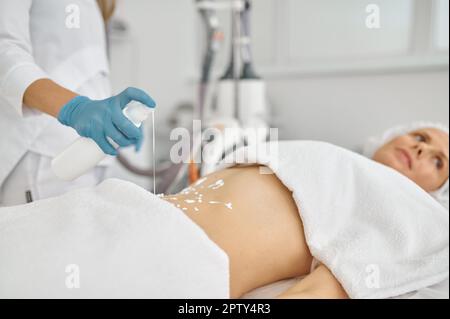 Cosmetologiste mettant de la crème sur le ventre du patient pour le massage gpl de gros plan. Rajeunissement et restauration de la peau dans une clinique moderne Banque D'Images