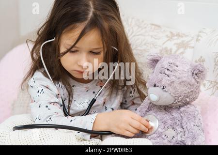 Grave, concentré, prudent petit enfant, fille vérifiant la poitrine de l'ours en peluche, respirer avec le stéthoscope, enveloppé dans une couverture. Santé et médecine. L Banque D'Images