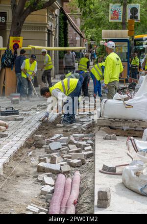 Les travailleurs qui pontent des pavés emboîtés pendant la construction du trottoir avec un sol surélevé rempli de tuyaux et de tuyaux à poser. Banque D'Images