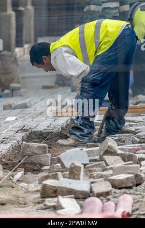 Les travailleurs qui pontent des pavés emboîtés pendant la construction du trottoir avec un sol surélevé rempli de tuyaux et de tuyaux à poser. Banque D'Images