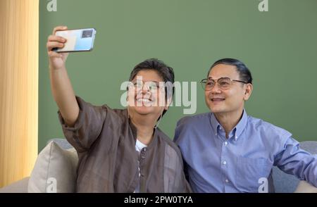 Un couple asiatique passe du temps ensemble dans le salon. Femme et homme à la retraite Prenez des photos de selfie avec un téléphone portable. Style de vie moderne personnes relation a Banque D'Images