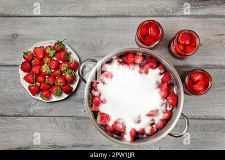 Vue sur table, grand pot de fraises en acier recouvert de cristaux de sucre, trois flacons avec fruits au sirop et la plaque à côté. La fraise fait maison Banque D'Images
