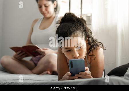 petite fille regardant curieusement son téléphone cellulaire, allongé sur le lit pendant que sa mère lit un livre. Banque D'Images