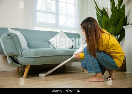 Femme de ménage nettoyage de la poussière sol sous canapé ou mobilier de canapé avec aspirateur, jeune femme asiatique heureuse avec accumulateur aspirateur à hom Banque D'Images