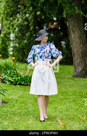 Portrait d'une fille souriante en chapeau noir à large bord, blouse bleue et jupe blanche debout sur l'herbe dans le parc. Banque D'Images