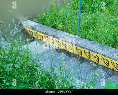 Indicateur de niveau d'eau avec marches en pierre et inscription noire sur fond jaune sur un remblai bordé d'herbe. Banque D'Images