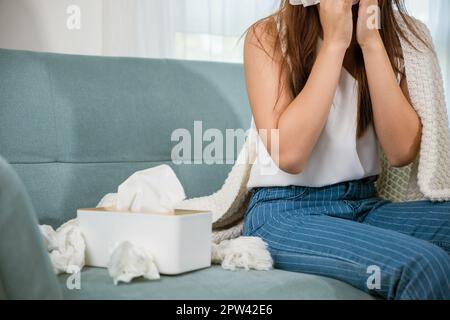 Malade femme assise sous une couverture sur le canapé et éternuer avec du papier de soie dans la salle de séjour, fièvre attrapée froid, asiatique jeune femme elle malade soufflant le nez snee Banque D'Images