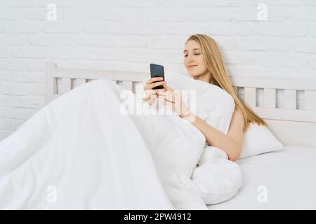Une jeune femme s'est réveillée et a commencé à envoyer des SMS sur un téléphone portable tout en étant au lit Banque D'Images