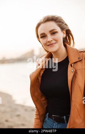 Bonne jeune femme dans une veste en cuir élégante souriante et élégante à la caméra tout en se tenant sur la côte du lac dans le vent soirée Banque D'Images