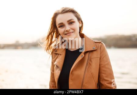 Bonne jeune femme dans une veste en cuir élégante souriante et élégante à la caméra tout en se tenant sur la côte du lac dans le vent soirée Banque D'Images