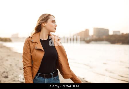 Bonne jeune femme dans une veste en cuir élégante souriante et élégante à la caméra tout en se tenant sur la côte du lac dans le vent soirée Banque D'Images