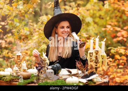 Jeune femme en costume de sorcière assise à table avec magie ingrédients et notes le jour de l'automne en forêt Banque D'Images