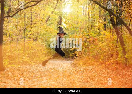 Femme en costume de sorcière volant sur le balai sur le chemin couvert de feuilles d'automne le jour d'Halloween dans la forêt Banque D'Images