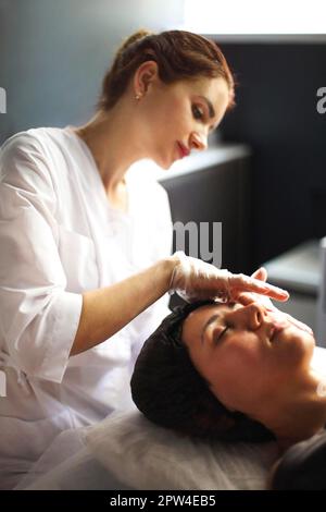 Cliché vertical d'une esthéticienne professionnelle en gants médicaux faisant un massage du visage pour une jeune femme au spa salon de beauté, massage et soin de la peau, femme Banque D'Images