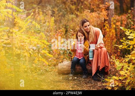 Jeune famille, mère et petite fille, dans des vêtements élégants, sirotant une boisson chaude tout en étant assis dans la forêt d'automne Banque D'Images