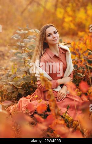 Belle jeune femme blonde portant une robe longue de style boho tout en étant assise avec un look pensif dans l'herbe haute en forêt d'automne décorée dans le rouge et le jaune Banque D'Images