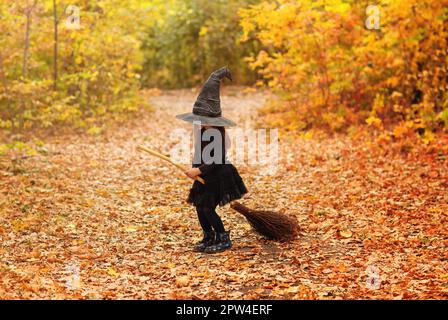 Mignonne heureuse petite fille à cheveux rouges vêtue d'un costume de sorcière assise sur un balai sur fond de forêt d'automne avec espace de copie, tenant son chapeau avec les mains. Banque D'Images