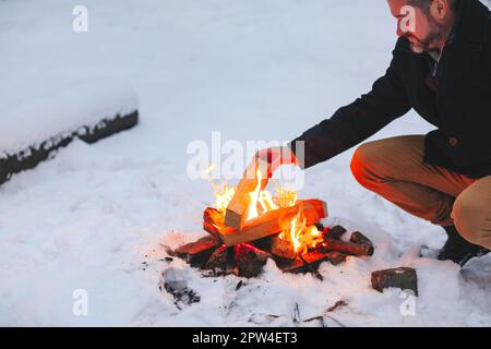 Homme mûr réchauffant ses mains avec le feu après avoir fait feu de camp au milieu de la forêt neigeuse en hiver froid soir, en utilisant la chaleur pour se sentir bien et Banque D'Images