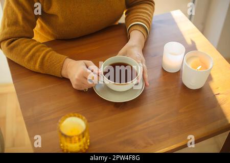 Photo de la femme avec une tasse de thé assis à la table avec des bougies allumées, femelle méconnaissable tenant une tasse de boisson chaude du matin pendant qu'elle est assise Banque D'Images