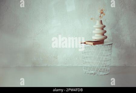 Livre et branche séchée dans un vase en céramique sur une table de chevet ronde en métal blanc contre un mur en béton beige clair dans un style loft simple intérieur d'appartement Banque D'Images