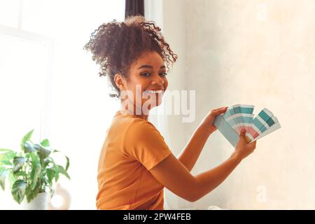 Souriante Afro-américaine fille se tient à l'appareil photo au milieu de la chambre avec la palette de nuances bleues pour la rénovation intérieure, choisir la peinture pour Banque D'Images