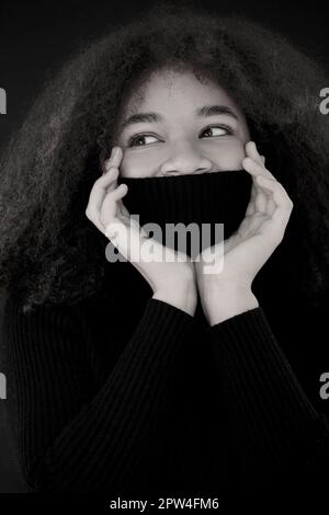 Heureuse afro-américaine curly femme fermer les yeux et couvrant le visage avec chaud noir col pull isolé sur fond sombre studio, souriante femme Banque D'Images