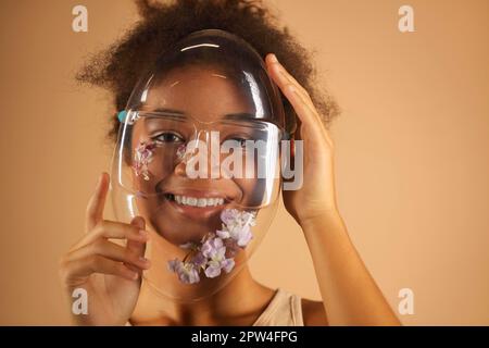 Jeune femme afro-américaine gaie portant un masque de protection en PVC transparent orné de fleurs et regardant la caméra avec des faisceaux Banque D'Images