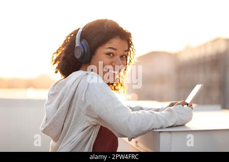 Portrait de côté d'une femme afro-américaine joyeuse dans un casque, écoutant de la musique sur un smartphone et souriant à l'appareil photo le matin Banque D'Images