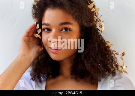 Gros plan portrait d'une femme afro-américaine mignonne avec des cheveux bouclés dans un serre-tête à feuilles d'or, avec un aspect flirté à l'appareil photo isolé sur blanc, jolie femme avec Banque D'Images