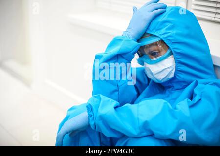 Femme fatiguée médecin dans l'équipement de protection individuelle EPI se sentant déprimé après un déplacement stressant difficile pendant la pandémie Covid-19, assis sur le sol Banque D'Images