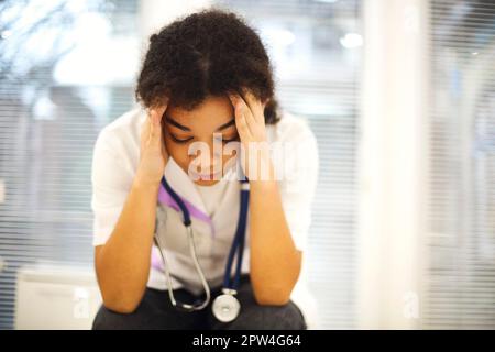 Triste déprimé jeune afro-américaine infirmière assis avec l'expression du visage frustré, se sentant submergé par la charge de travail dans la clinique, ethnique médecin féminin Banque D'Images