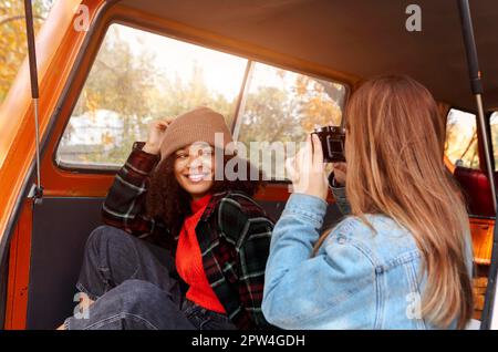 Jeune femme afro-américaine souriante posant pour la photo tout en étant assise sur le coffre de voiture avec une amie féminine, deux amies multiraciales diverses en monospace Banque D'Images