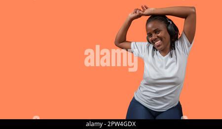 Portrait d'une jeune femme africaine heureuse dansant et écoutant de la musique avec des écouteurs, des bras vers le haut, vêtue d'un t-shirt blanc et d'un pantalon bleu contre l'orange Banque D'Images