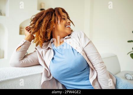 Jeune fille millénaire afro-américaine, heureuse et heureuse, asseyez-vous sur un canapé confortable dans un salon moderne et confortable. Profitez de sa vie Banque D'Images